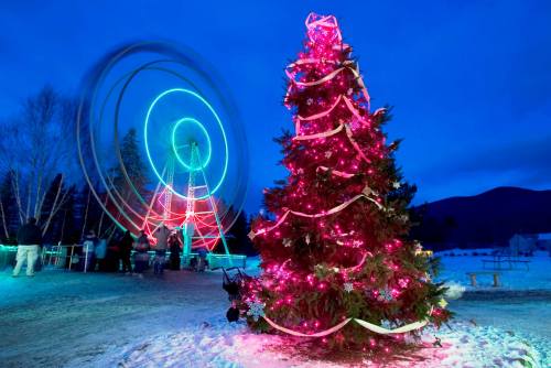 A brightly lit Christmas tree in the foreground with a colorful ferris wheel spinning in the background at dusk.