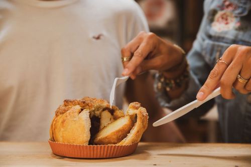 Two hands using plastic knives to slice a round, crumbly cake on a wooden table.