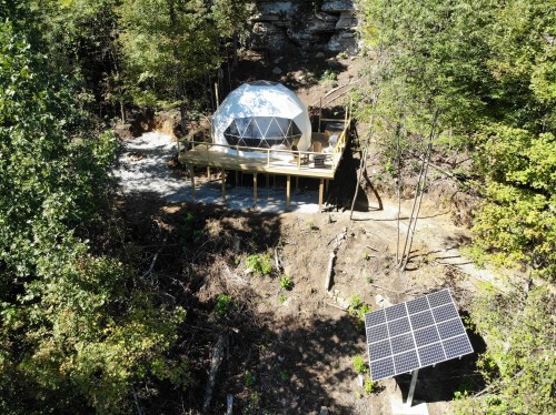 A geodesic dome home on a wooden deck surrounded by trees, with a solar panel nearby.