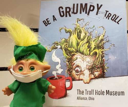 A troll doll in a mask stands in front of a sign for the Troll Hole Museum, featuring a grumpy troll illustration.