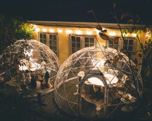 Two transparent igloo-shaped structures with tables inside, illuminated by string lights, set in a cozy outdoor space at night.