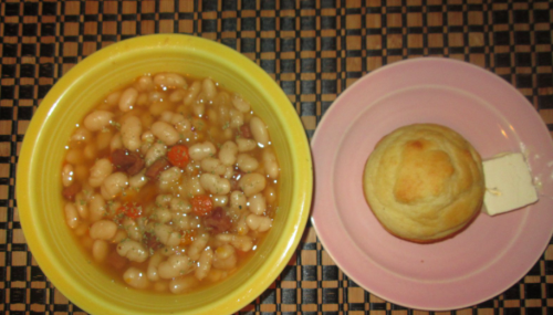 A bowl of white bean soup with herbs and a pink plate holding a cornbread muffin with a pat of butter.