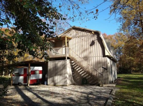 A two-story wooden house with a staircase, surrounded by trees and a gravel driveway.