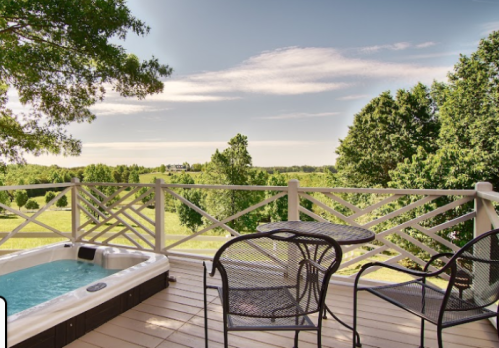 A scenic view from a deck featuring a hot tub, two chairs, and a table, surrounded by lush greenery and a clear sky.