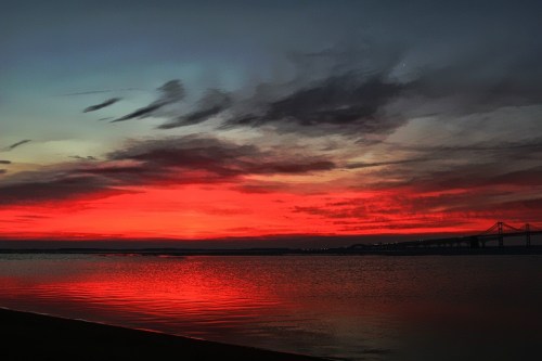 A vibrant sunset with red and orange hues reflecting on calm water, silhouetting a bridge in the background.