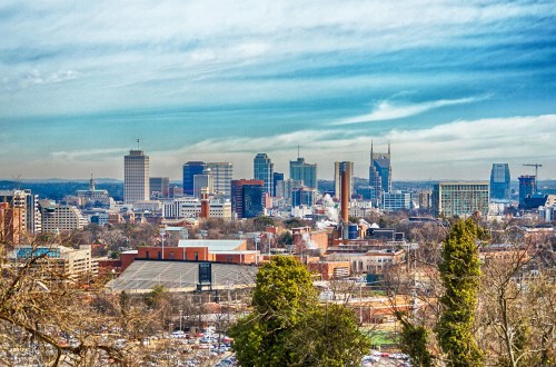 A panoramic view of a city skyline with tall buildings, trees in the foreground, and a cloudy sky above.
