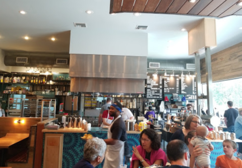 A busy restaurant interior with patrons dining, staff working in the open kitchen, and a menu board in the background.