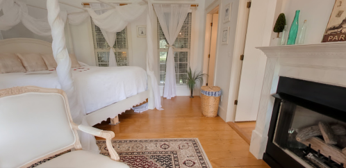 Cozy bedroom with a four-poster bed, white drapes, a fireplace, and a decorative rug on wooden floors.