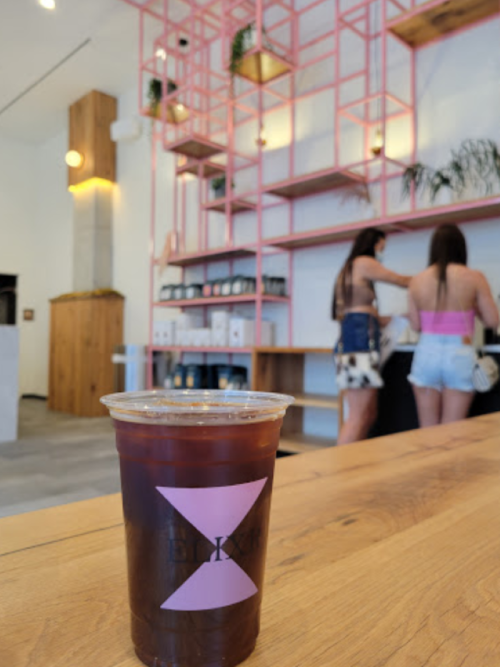 A clear cup of iced coffee sits on a wooden table, with two people preparing drinks in the background.