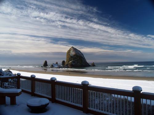 Snow-covered beach with a rocky outcrop, waves gently lapping at the shore under a cloudy sky.
