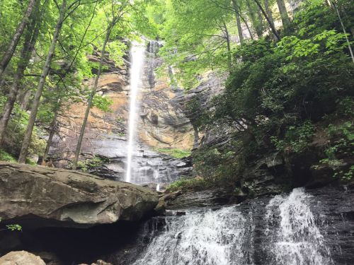 A tall waterfall cascades down rocky cliffs, surrounded by lush green trees and a flowing stream at the base.