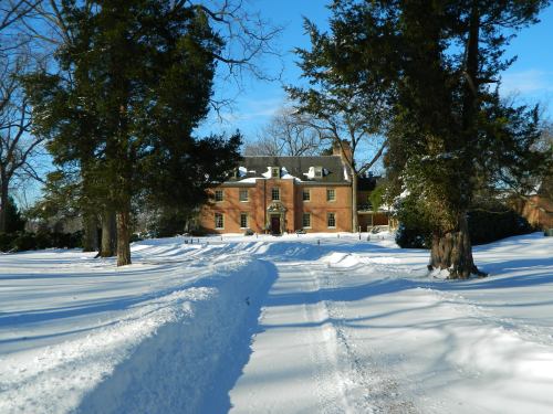 A snow-covered path leads to a large brick house surrounded by trees on a clear winter day.