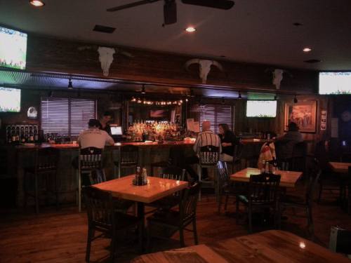 A rustic bar interior with wooden tables, a long bar, and patrons seated at the bar, with TVs showing sports.