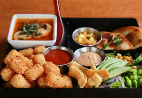 A black tray filled with crispy tater tots, dipping sauces, a bowl of soup, and assorted vegetables and sides.