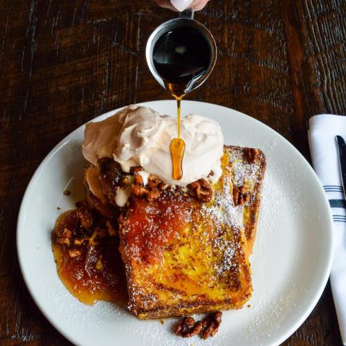 A plate of French toast topped with whipped cream, nuts, and syrup, served on a wooden table.
