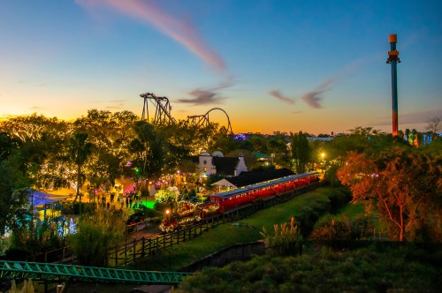 A vibrant amusement park at sunset, featuring a train, roller coasters, and colorful lights among lush trees.
