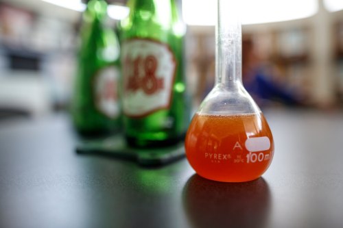 A Pyrex flask with orange liquid sits on a table, with green bottles blurred in the background.