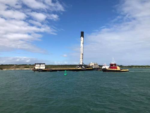 A barge with a tall rocket booster is anchored in the water, with tugboats nearby and a clear blue sky above.