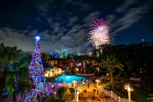 A festive scene with a decorated Christmas tree, a pool, and fireworks lighting up the night sky at an amusement park.