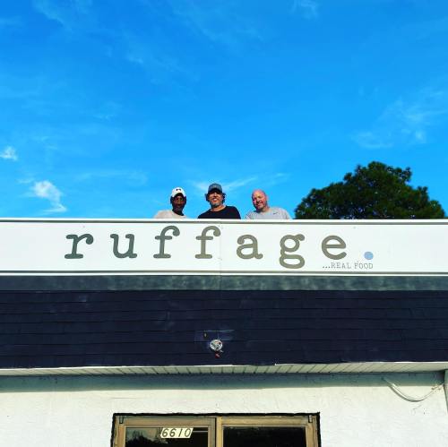 Three men stand on the roof of a building with a sign that reads "ruffage ...REAL FOOD" against a blue sky.