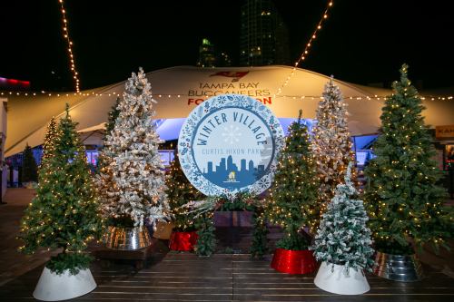 A festive scene with decorated Christmas trees and a sign for "Winter Village" at Curtis Hixon Park, Tampa.