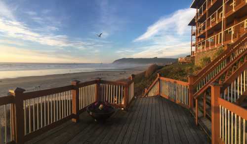 A wooden deck overlooks a beach at sunrise, with a seagull flying and cliffs in the distance.