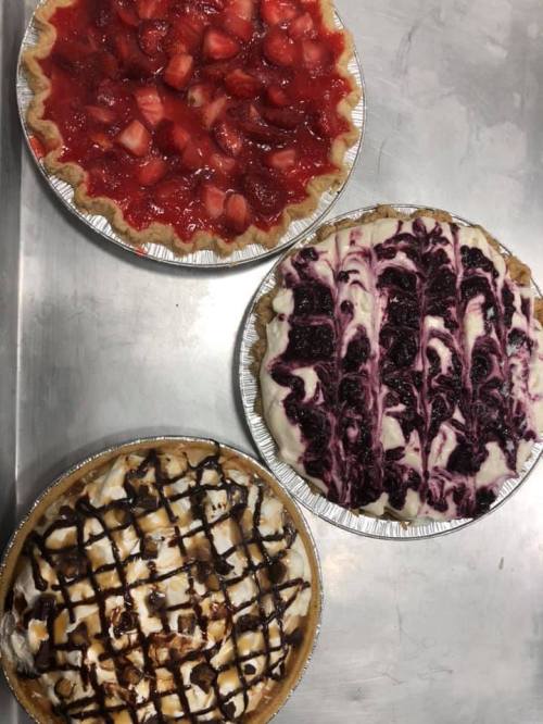 Three delicious pies on a silver tray: a strawberry pie, a blueberry cream pie, and a chocolate-drizzled pie.