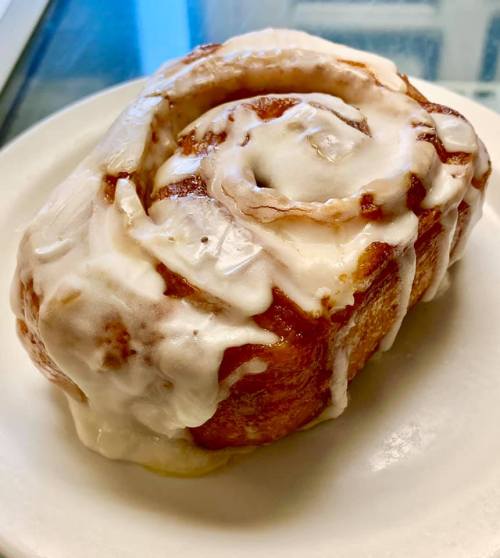 A freshly baked cinnamon roll topped with creamy icing, sitting on a white plate.