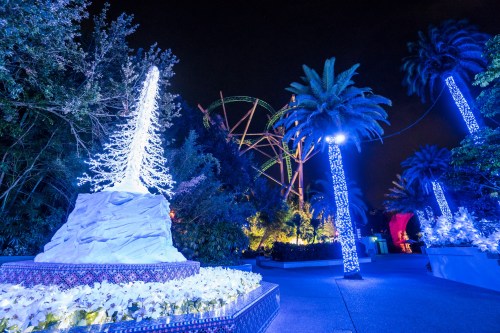 A festive scene with a lit Christmas tree, palm trees, and colorful lights at night, with a roller coaster in the background.