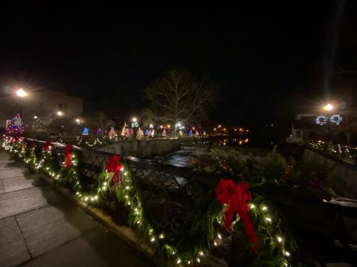 A festive nighttime scene by a river, adorned with colorful lights and holiday decorations along a walkway.