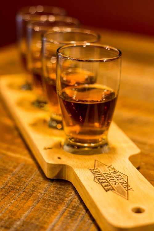 A wooden flight board holds four glasses of amber liquid, set on a rustic wooden table.