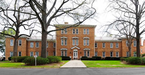 A large brick building with multiple windows, surrounded by trees and a manicured lawn.
