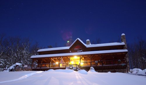 A cozy cabin illuminated at night, surrounded by snow and trees under a starry sky.
