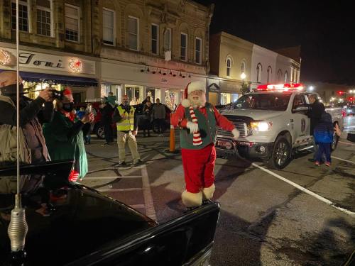 A festive scene with Santa Claus waving at a nighttime parade, surrounded by onlookers and emergency vehicles.