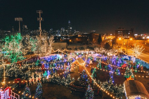Aerial view of a festive park decorated with colorful Christmas lights and trees, with a city skyline in the background.