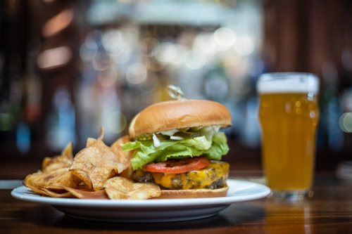 A juicy cheeseburger with lettuce, tomato, and pickles, served with chips and a glass of beer in a bar setting.