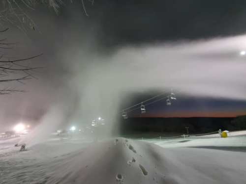 A snowy ski slope at night, with snowmaking machines creating mist and ski lifts in the background.