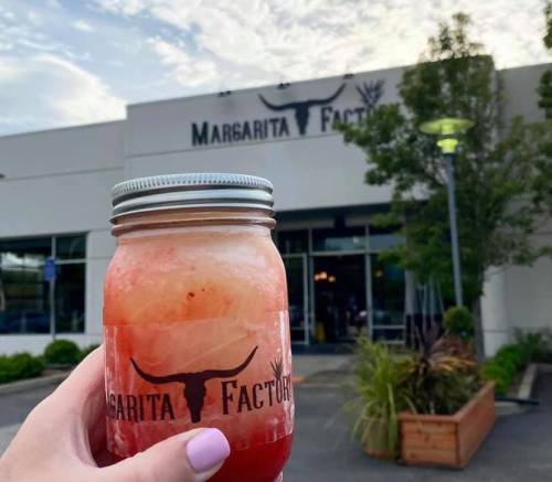A hand holding a drink in a mason jar in front of the Margarita Factory building under a cloudy sky.