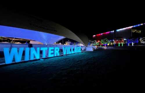 Brightly lit "WINTER VILLAGE" sign at night, surrounded by colorful holiday lights and festive decorations.