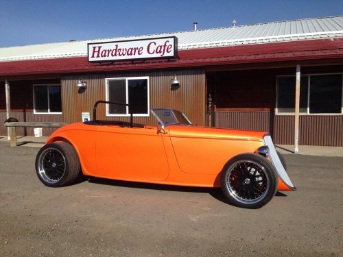 A bright orange vintage convertible car parked in front of a rustic cafe with a sign that reads "Hardware Cafe."