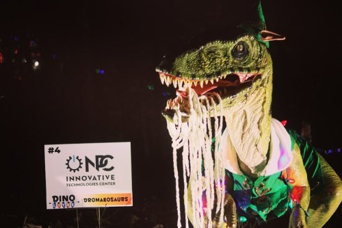 A dinosaur costume with colorful lights, wearing a festive outfit, stands next to a sign for an innovative technology center.