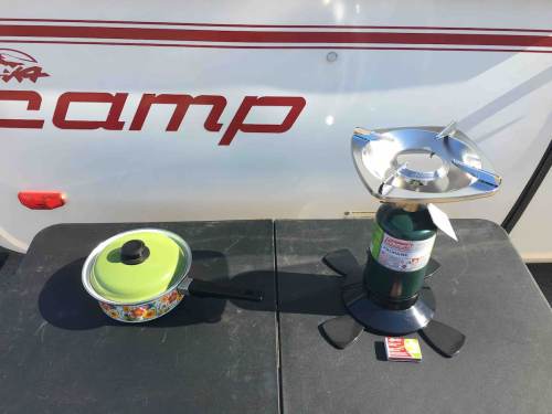 A camping stove next to a pot with a lid on a black table, with a camper in the background.