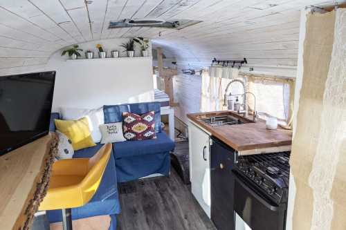 Cozy interior of a converted bus, featuring a kitchen, seating area, and bright decor with plants and cushions.