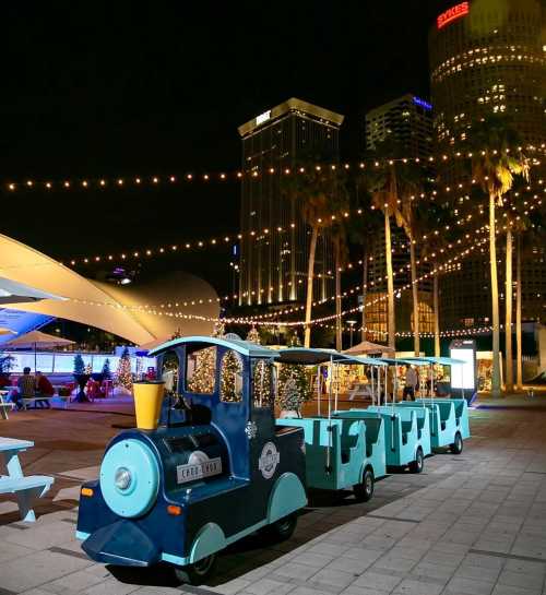 A colorful train-shaped vehicle parked at night, surrounded by palm trees and city buildings, with string lights overhead.