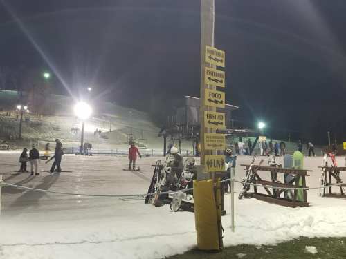 A snowy ski area at night with people skiing, signs for amenities, and picnic tables in the foreground.