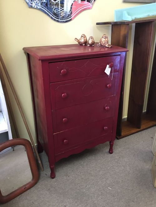 A red wooden chest of drawers with four drawers, placed in a room with other furniture. Two decorative birds on top.