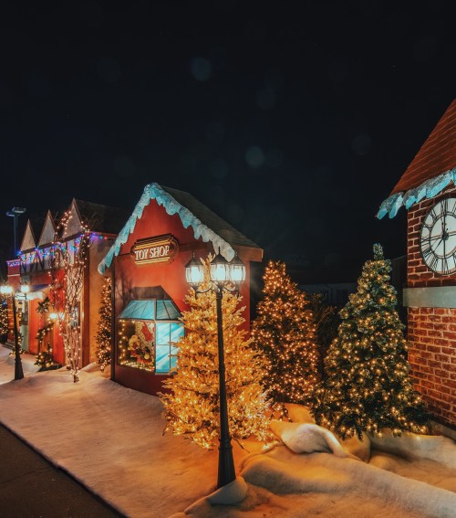 A festive scene featuring a toy shop, decorated trees, and snow, illuminated by colorful lights at night.