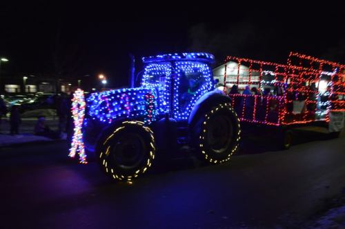 A brightly lit tractor decorated with colorful lights pulls a festive trailer during a nighttime parade.