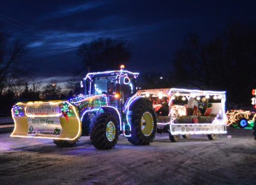 A brightly lit tractor with a snowplow, decorated with colorful lights, against a dark evening sky.