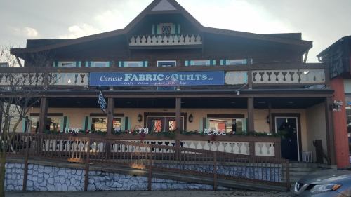 A large, wooden building with a sign reading "Carlisle Fabric & Quilts," featuring a welcoming porch and decorative elements.